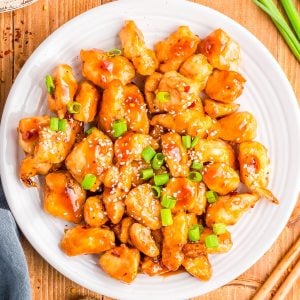 A white plate of orange chicken garnished with sesame seeds and chopped green onions, placed on a wooden table with ginger, green onions, and rice nearby.