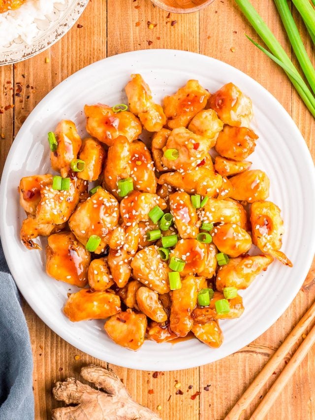 A white plate of orange chicken garnished with sesame seeds and chopped green onions, placed on a wooden table with ginger, green onions, and rice nearby.