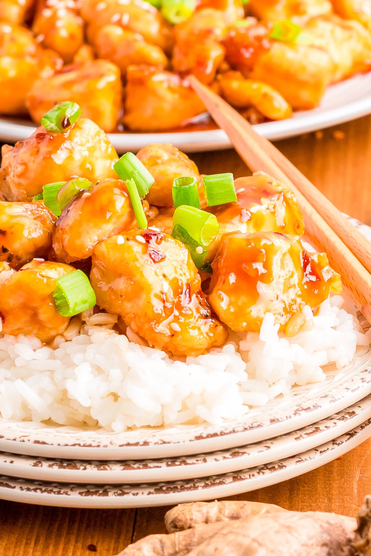 A plate of white rice topped with pieces of glazed orange chicken garnished with chopped green onions, with chopsticks resting on the side.