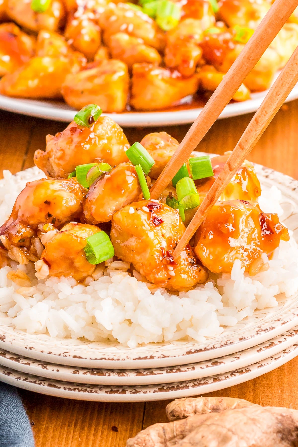 Chopsticks pick up pieces of glazed chicken garnished with green onions, served over white rice on a stacked plate.