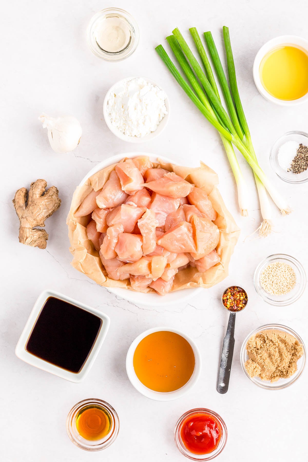 Overhead view of cubed raw chicken surrounded by various ingredients including soy sauce, honey, ginger, green onions, flour, oil, garlic, brown sugar, vinegar, and seasonings.