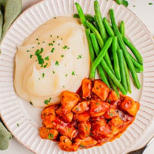 A plate with mashed potatoes, green beans, and barbecue-glazed chicken pieces, garnished with chopped parsley.