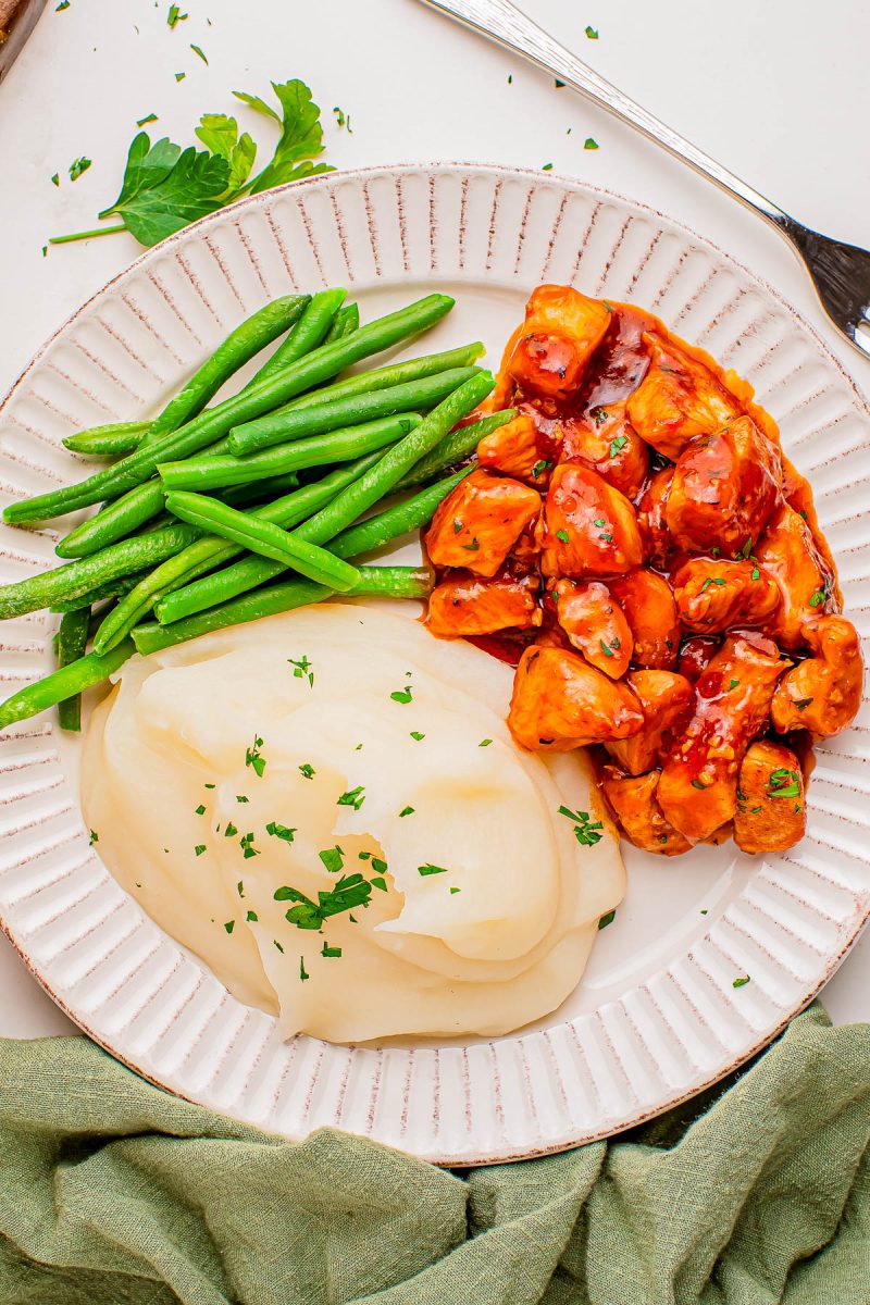 A plate with green beans, mashed potatoes topped with herbs, and diced chicken in a red sauce.