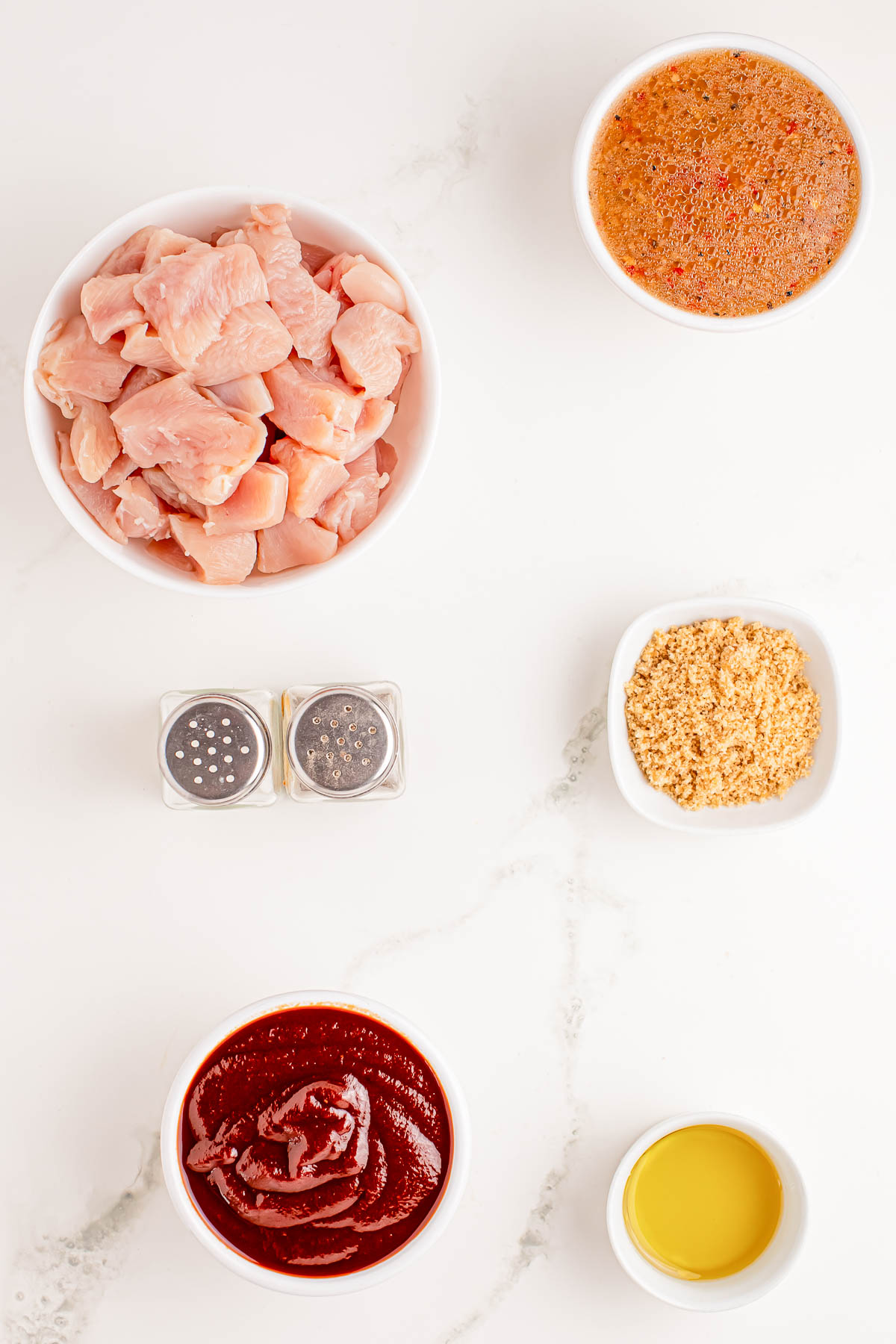 Overhead view of raw chicken pieces, barbecue sauce, marinade, minced garlic, olive oil, and salt and pepper on a white surface.