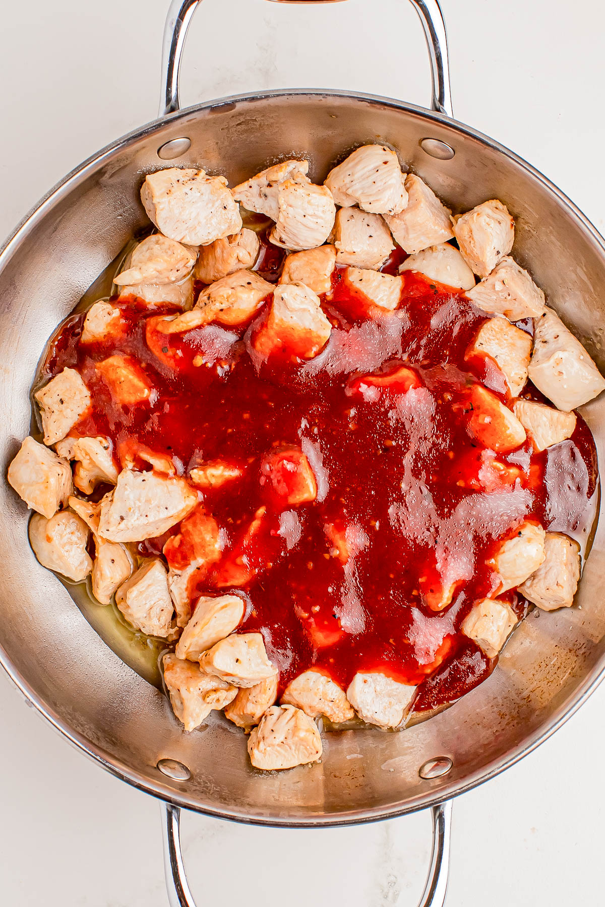 Chunks of cooked chicken in a metal pan topped with a layer of red sauce, ready to be mixed or cooked further.