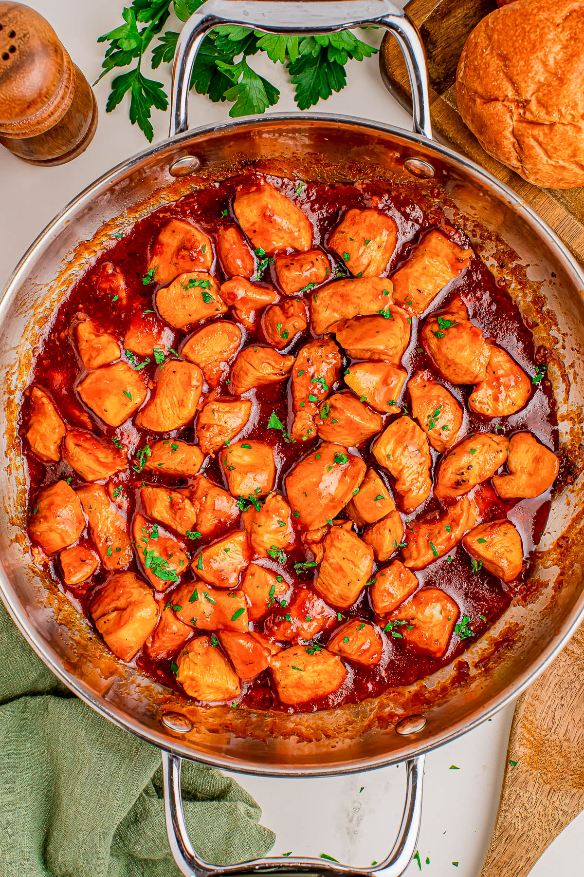 A stainless steel skillet filled with cooked, saucy chicken pieces garnished with chopped herbs, with bread, parsley, and a pepper mill nearby.