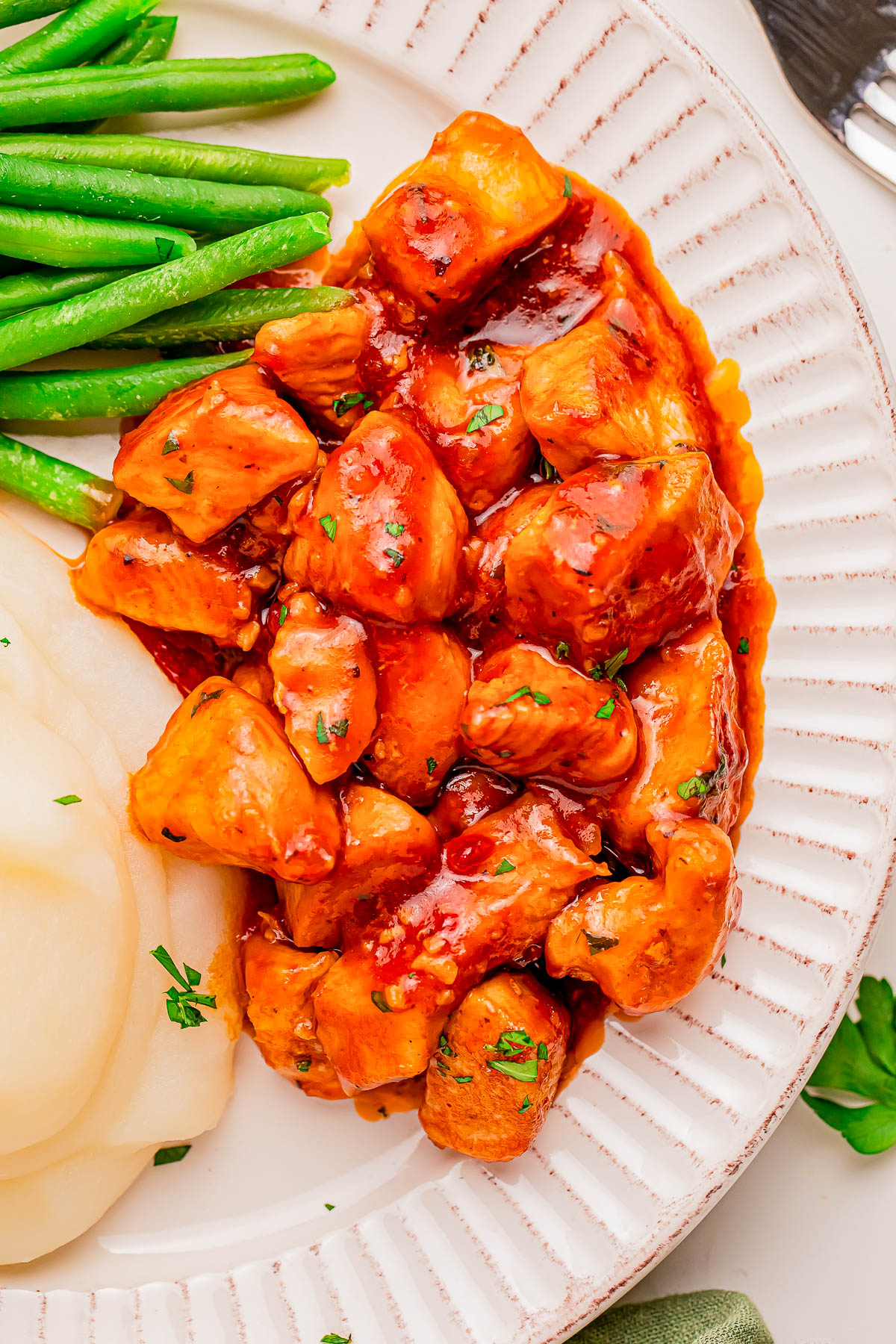 Plate of diced chicken in a reddish sauce, served with mashed potatoes and green beans on a white patterned plate.