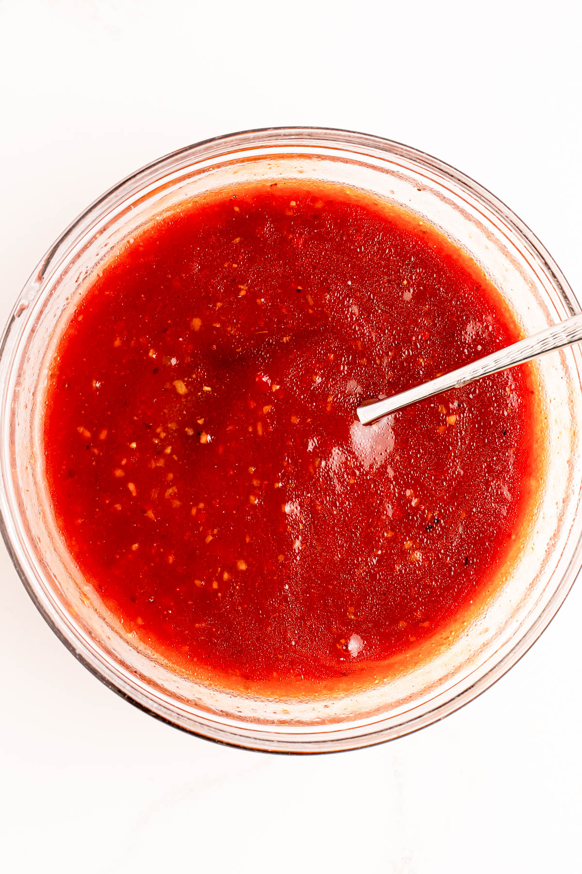 A bowl of red salsa with visible seasoning and a spoon resting in it, viewed from above on a white surface.