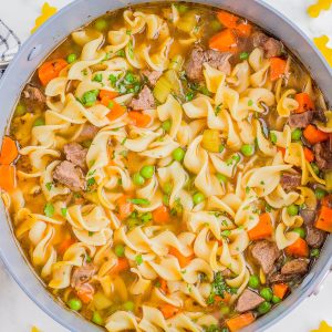 A pot of beef noodle soup with egg noodles, carrots, peas, celery, and chunks of beef, garnished with parsley. Uncooked pasta and herbs are scattered on the white surface nearby.
