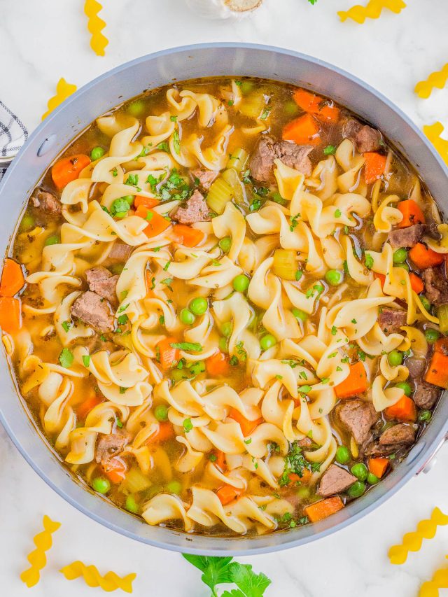 A pot of beef noodle soup with egg noodles, carrots, peas, celery, and chunks of beef, garnished with parsley. Uncooked pasta and herbs are scattered on the white surface nearby.