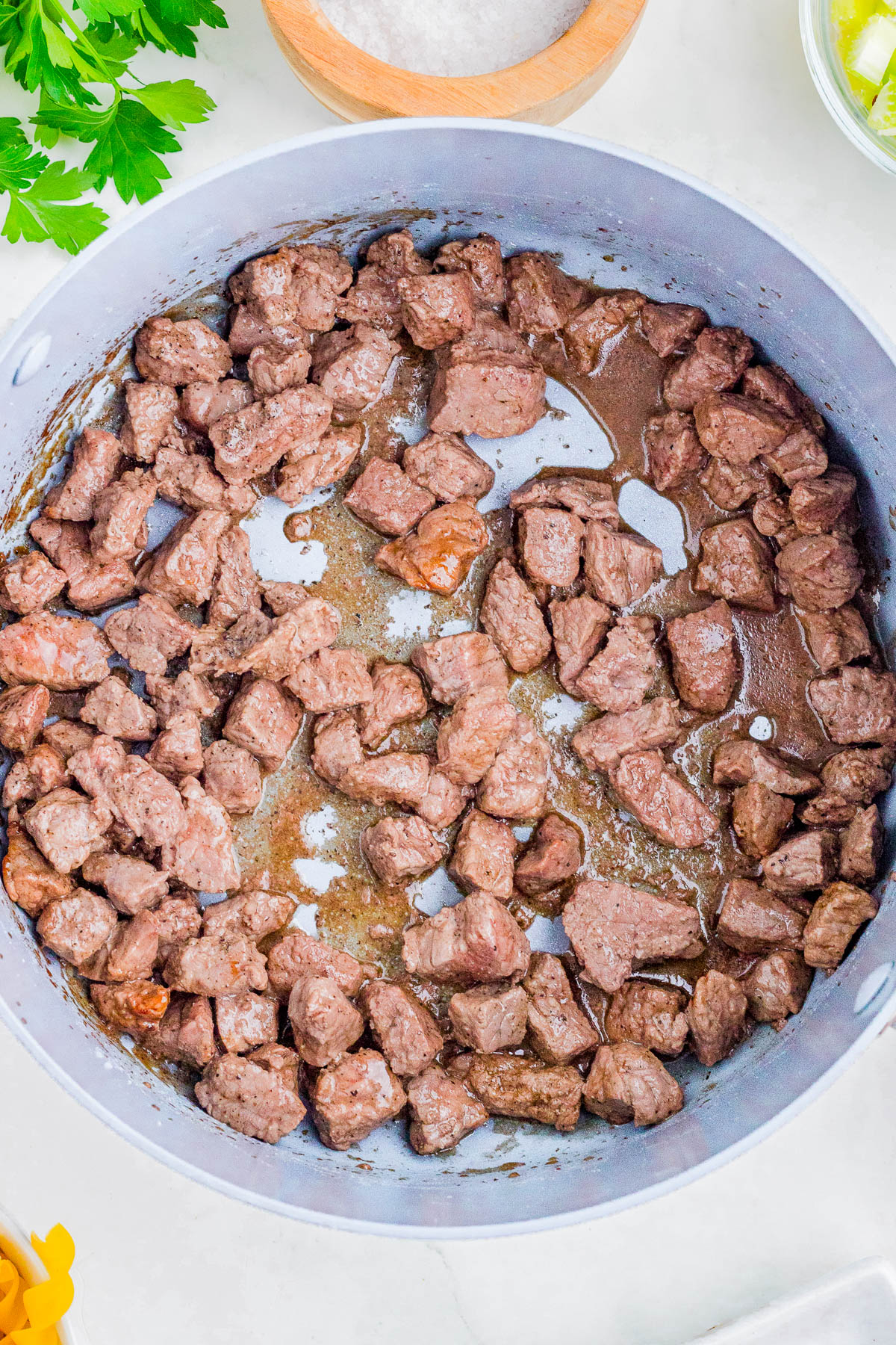 Cubed pieces of beef are browning in a large gray skillet with some liquid at the bottom. Fresh parsley and other ingredients are visible around the skillet.