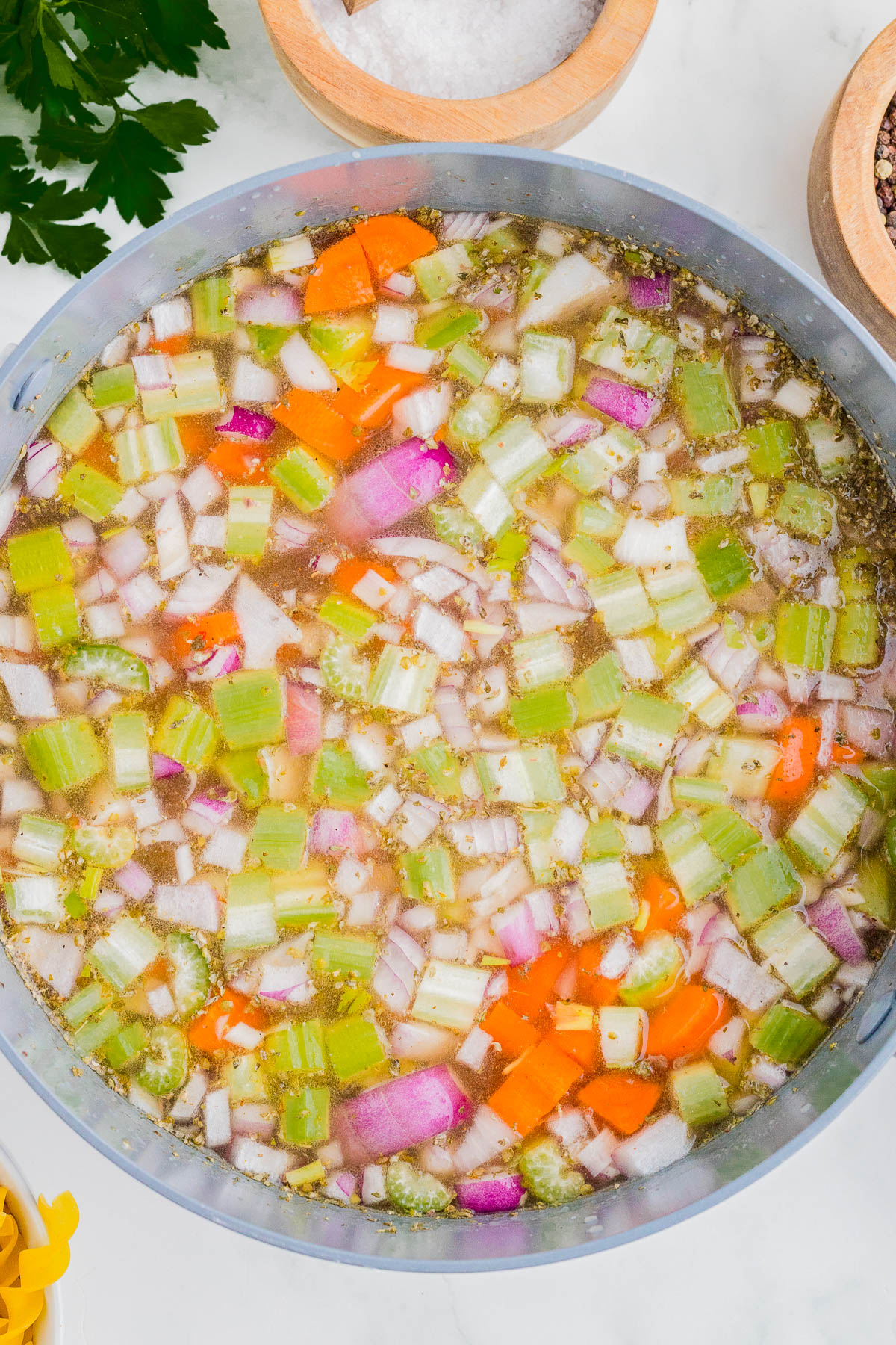 A pot filled with chopped celery, carrots, red onions, and seasoning in broth, ready for cooking on a white countertop.
