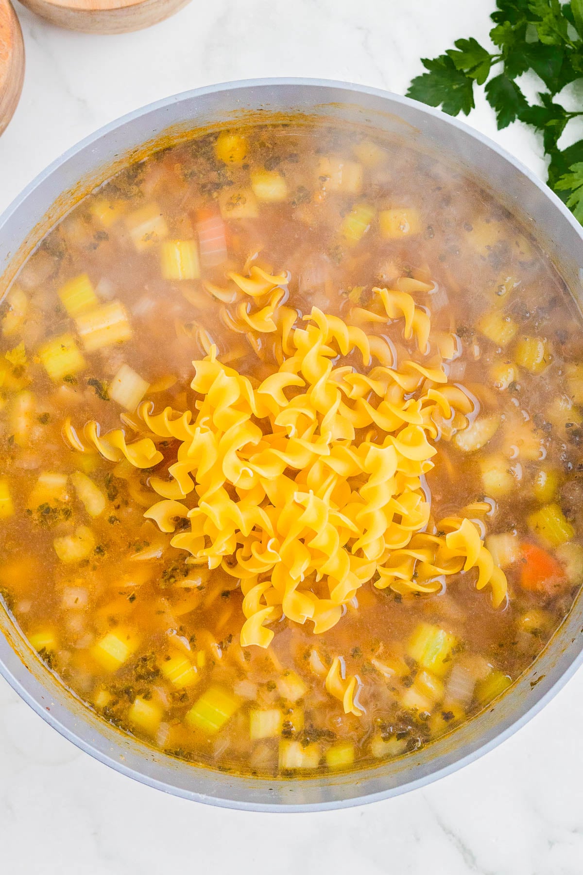 A pot of soup with chopped vegetables and uncooked egg noodles being added, with parsley and wooden bowls visible in the background.