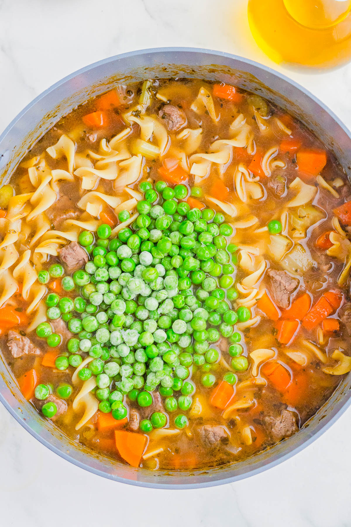 A pot of noodle soup with beef, carrots, and broth, topped with a pile of frozen green peas before being stirred in.
