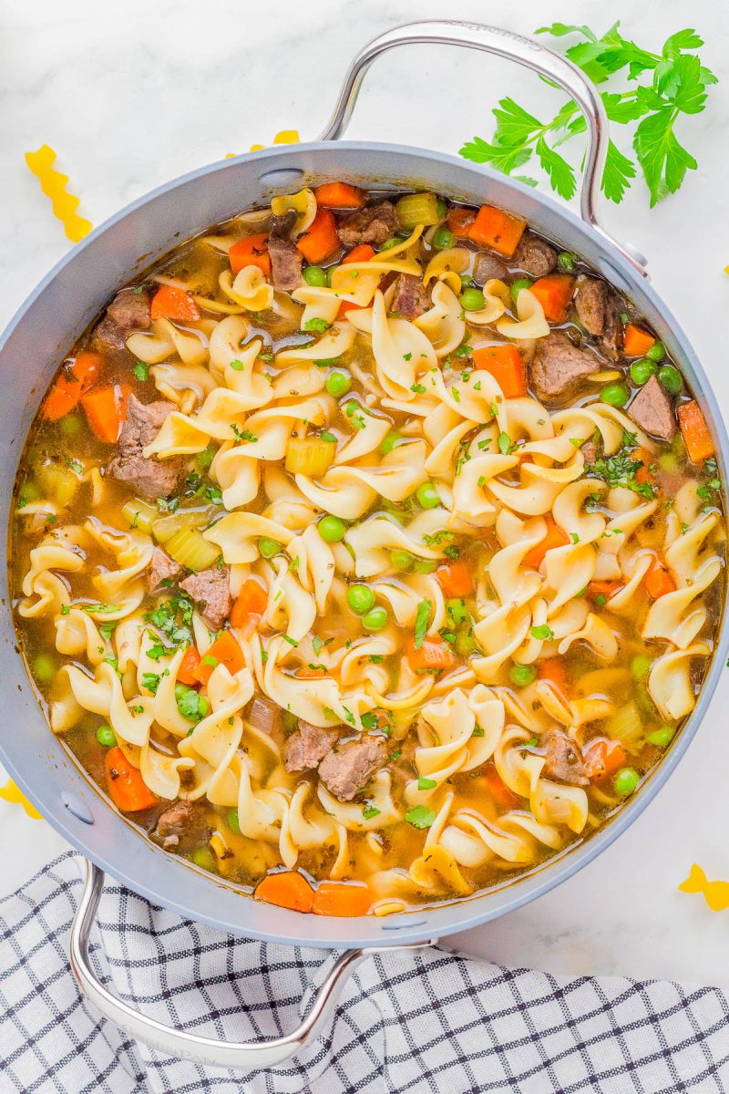 A pot of beef noodle soup with carrots, celery, peas, and parsley, featuring wide egg noodles in a clear broth. A checkered towel and fresh herbs are nearby.