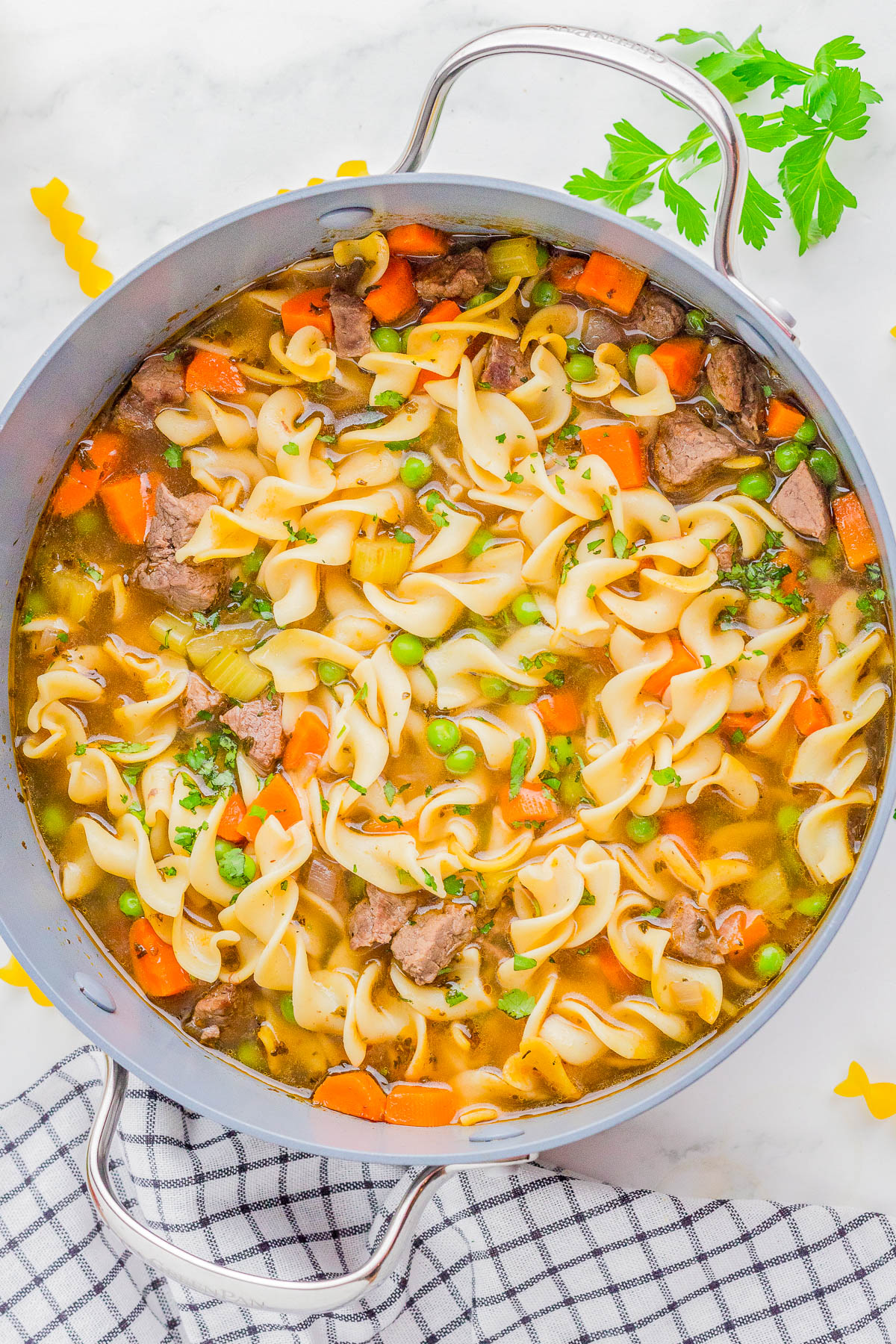 A pot of beef noodle soup with carrots, celery, peas, and parsley, featuring wide egg noodles in a clear broth. A checkered towel and fresh herbs are nearby.
