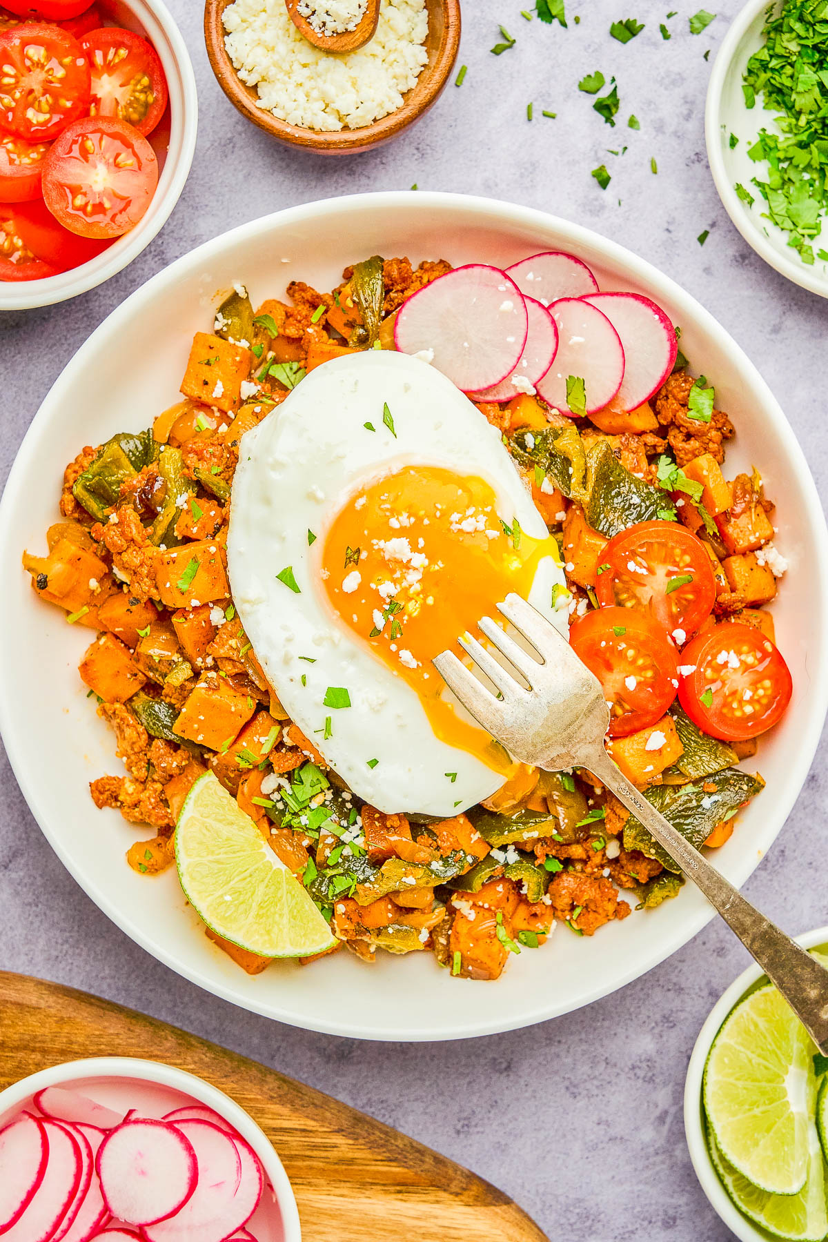 A bowl of diced vegetables and ground meat topped with a fried egg, sliced radishes, cherry tomatoes, lime wedge, and crumbled cheese, with a fork breaking the egg yolk.