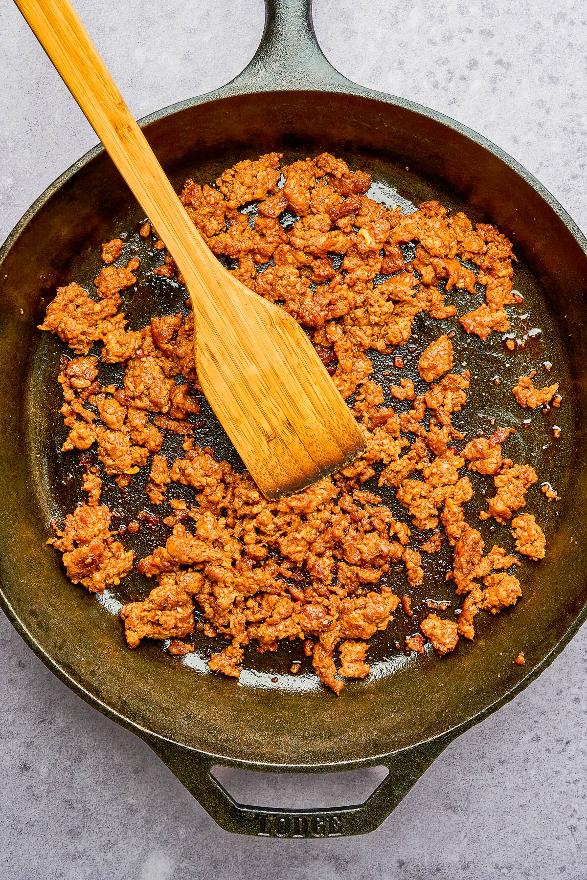 Cooked ground meat mixed with spices in a cast iron skillet, being stirred with a wooden spatula on a light-colored surface.