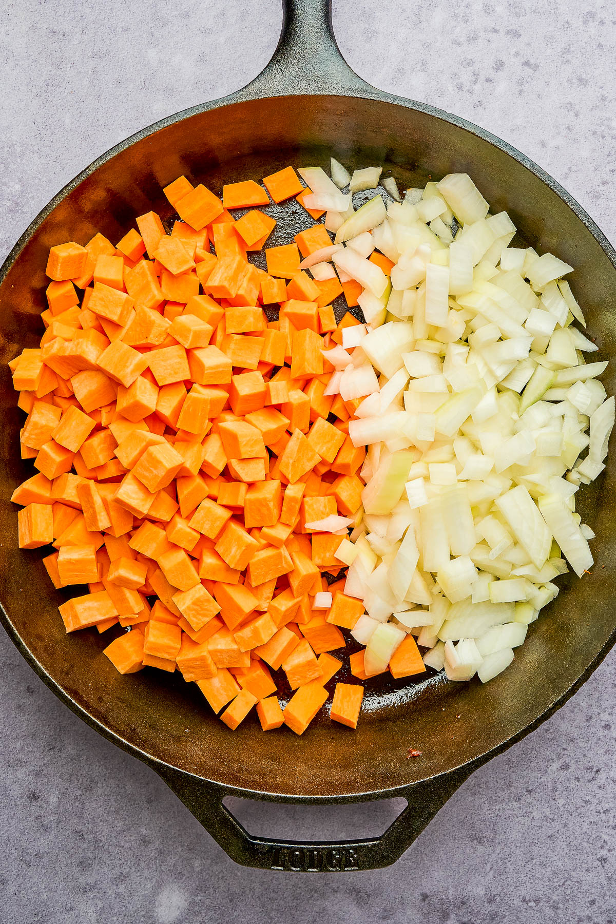 A cast iron skillet containing diced sweet potatoes on one side and diced onions on the other, placed on a gray surface.