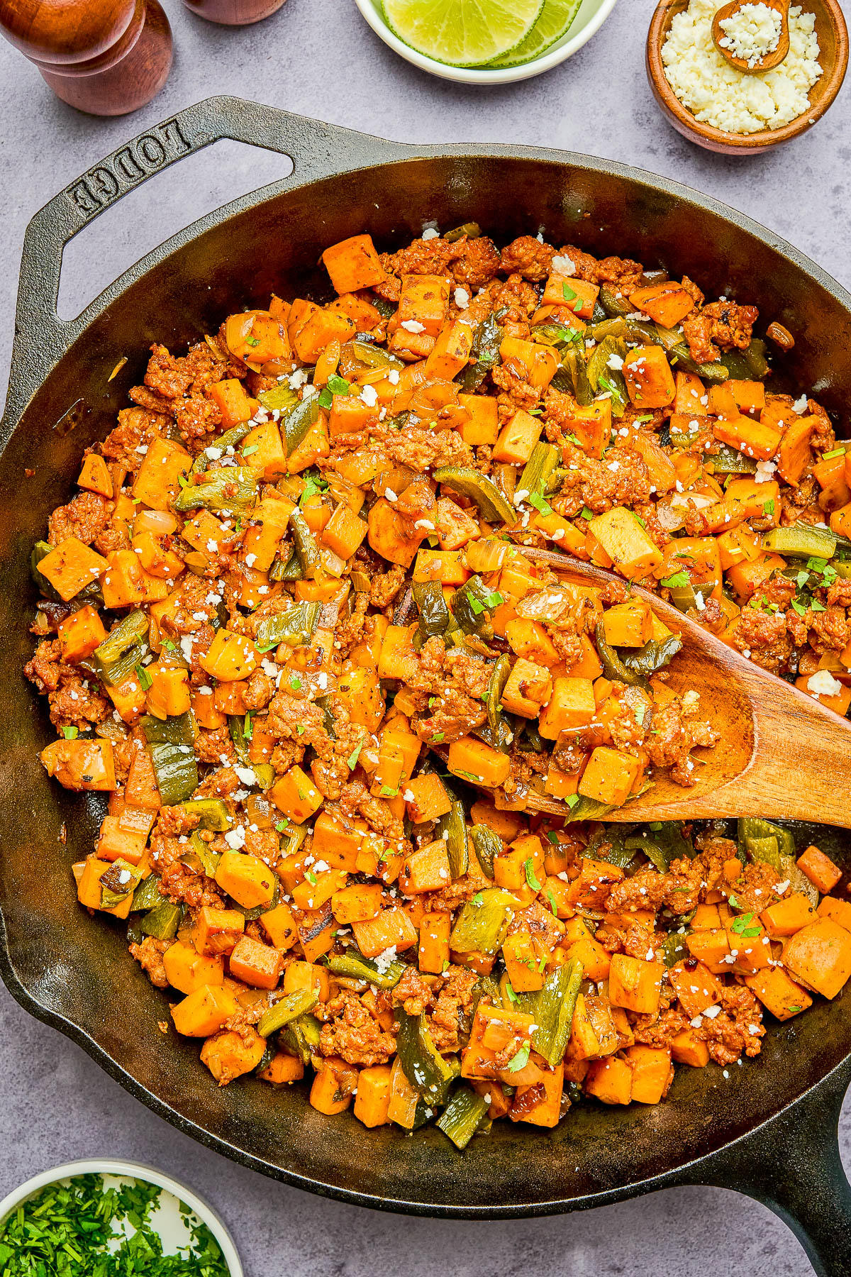 A cast iron skillet filled with a cooked mixture of ground meat, diced sweet potatoes, green bell peppers, and crumbled cheese, with a wooden spoon resting inside.