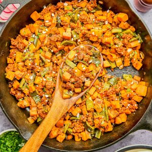A cast-iron skillet filled with a cooked mixture of ground meat, diced sweet potatoes, and green beans, with a wooden spoon resting inside. Lime wedges and eggs are shown on the side.