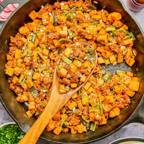 A cast-iron skillet filled with a cooked mixture of ground meat, diced sweet potatoes, and green beans, with a wooden spoon resting inside. Lime wedges and eggs are shown on the side.