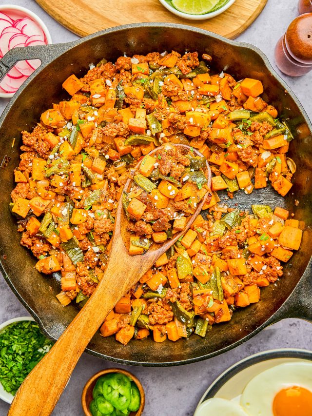 A cast-iron skillet filled with a cooked mixture of ground meat, diced sweet potatoes, and green beans, with a wooden spoon resting inside. Lime wedges and eggs are shown on the side.