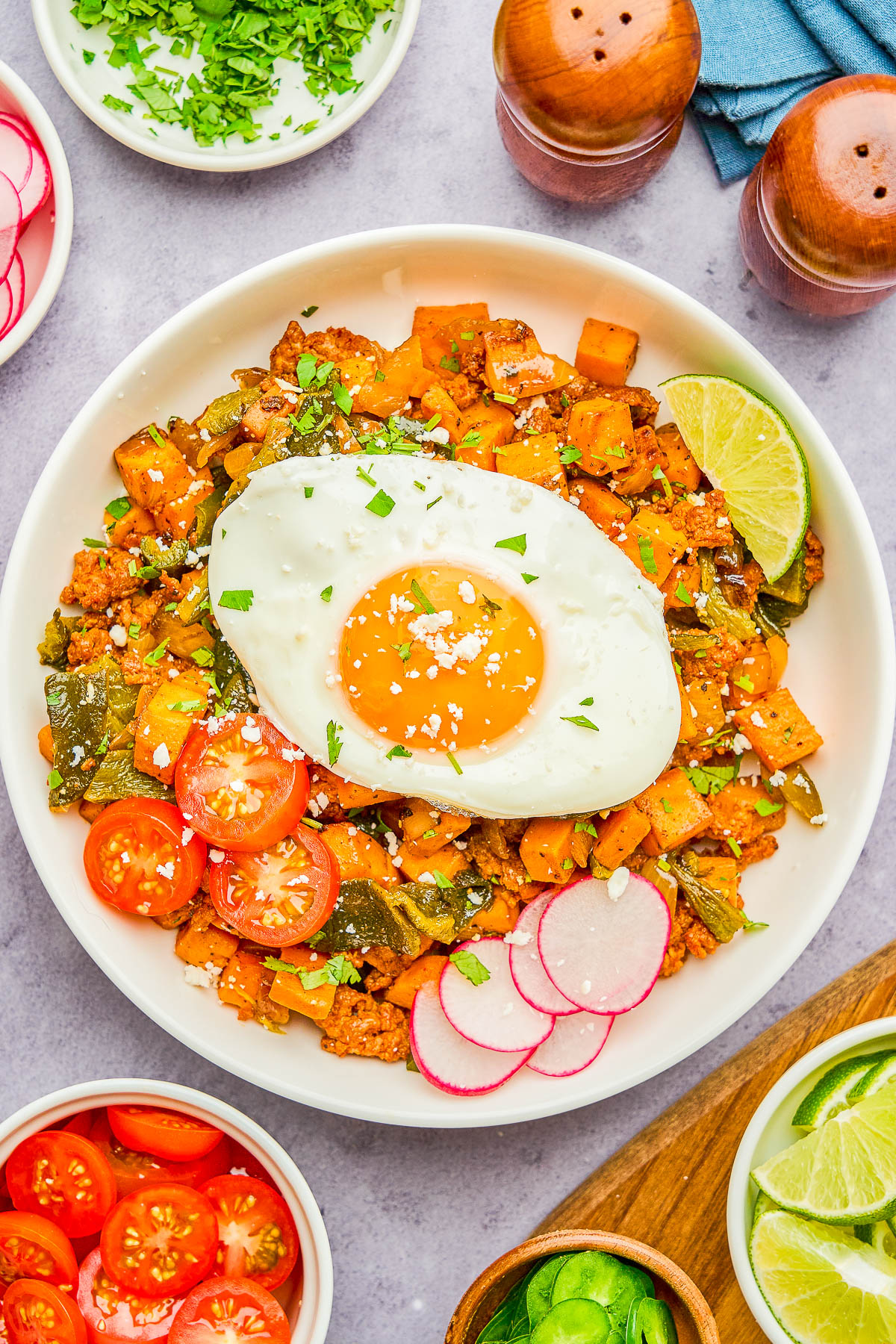 A bowl of hash with diced sweet potatoes, vegetables, and ground meat, topped with a sunny-side-up egg, sliced radishes, cherry tomatoes, and a lime wedge.