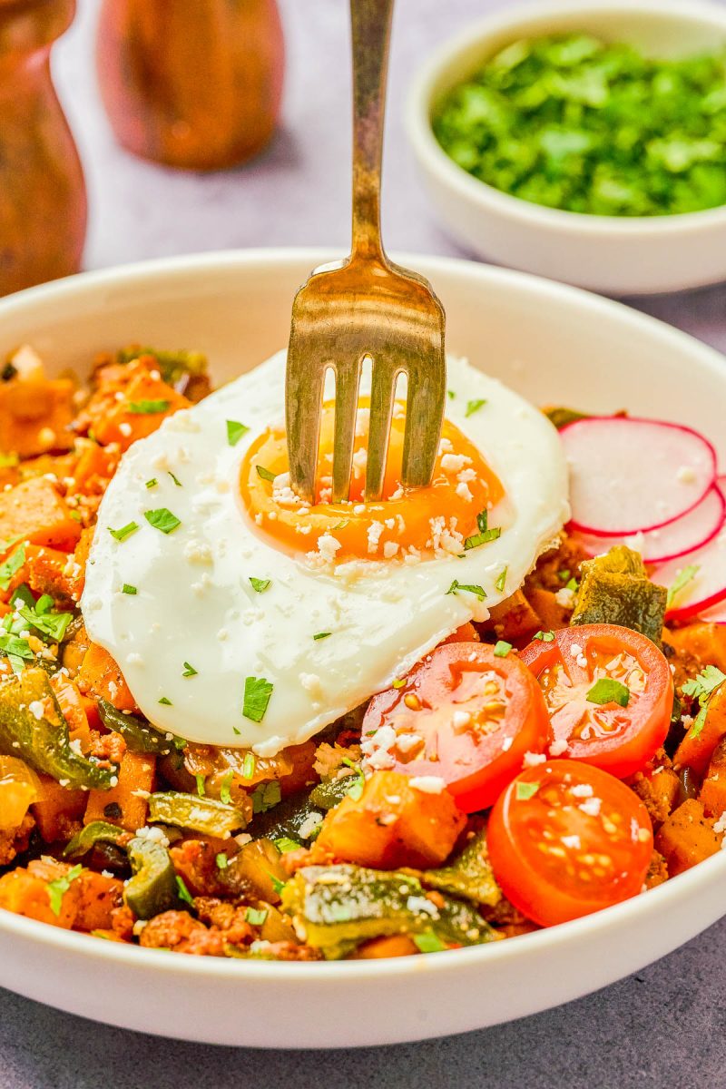 A fork is piercing the yolk of a sunny-side up egg on top of a vegetable hash with sliced cherry tomatoes and radishes in a white bowl, garnished with chopped herbs.