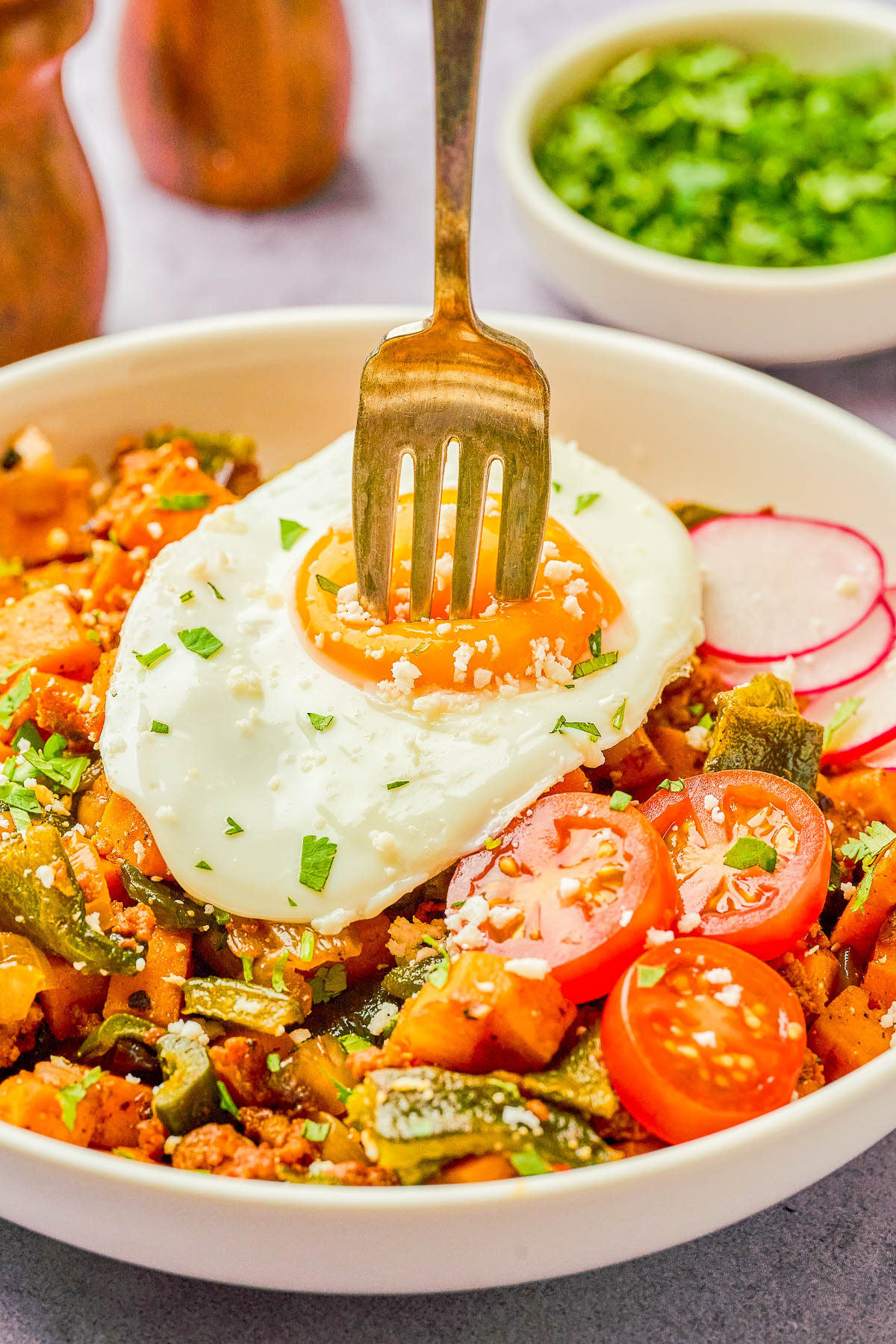 A fork is piercing the yolk of a sunny-side up egg on top of a vegetable hash with sliced cherry tomatoes and radishes in a white bowl, garnished with chopped herbs.