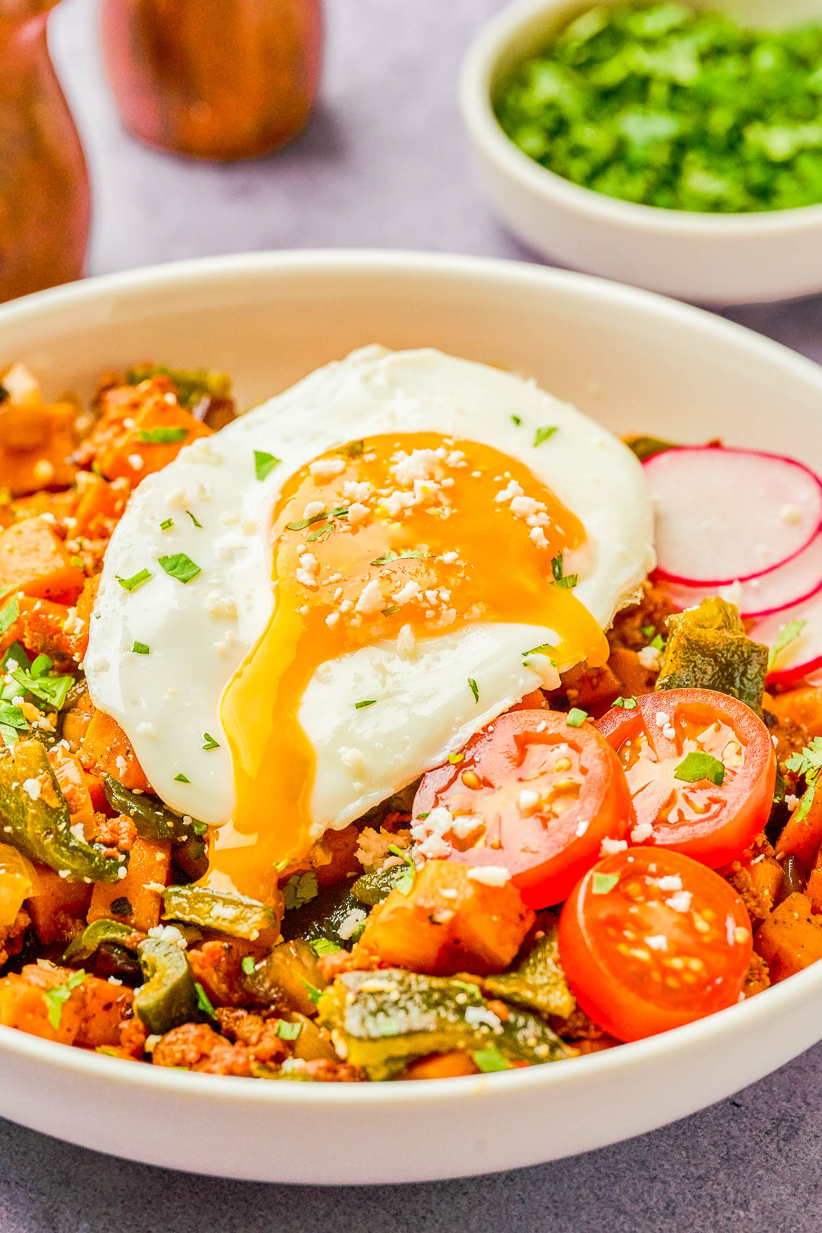 A bowl of hash topped with a runny fried egg, sliced cherry tomatoes, radish, and chopped herbs, with a small bowl of herbs in the background.