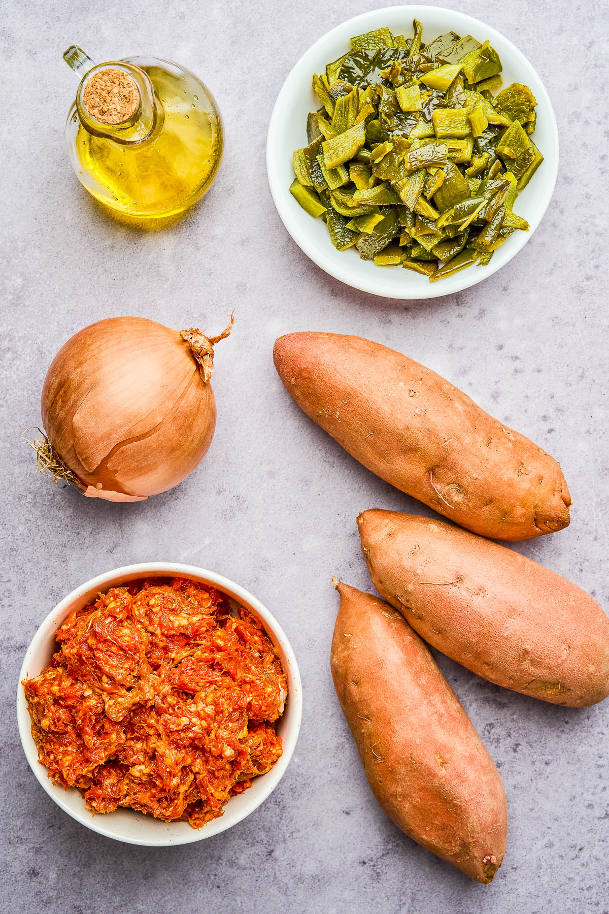 A bottle of olive oil, a whole onion, cooked nopales, raw sweet potatoes, and a bowl of seasoned ground meat are arranged on a light surface.