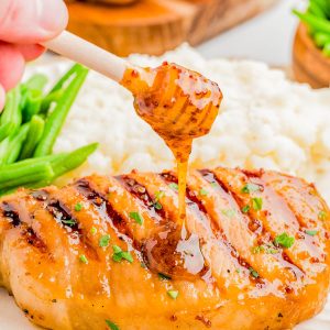 A hand drizzles honey mustard sauce onto a grilled chicken breast, served with mashed potatoes and green beans on a white plate.