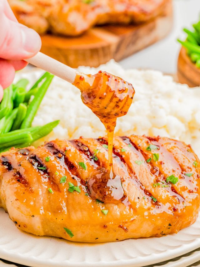 A hand drizzles honey mustard sauce onto a grilled chicken breast, served with mashed potatoes and green beans on a white plate.