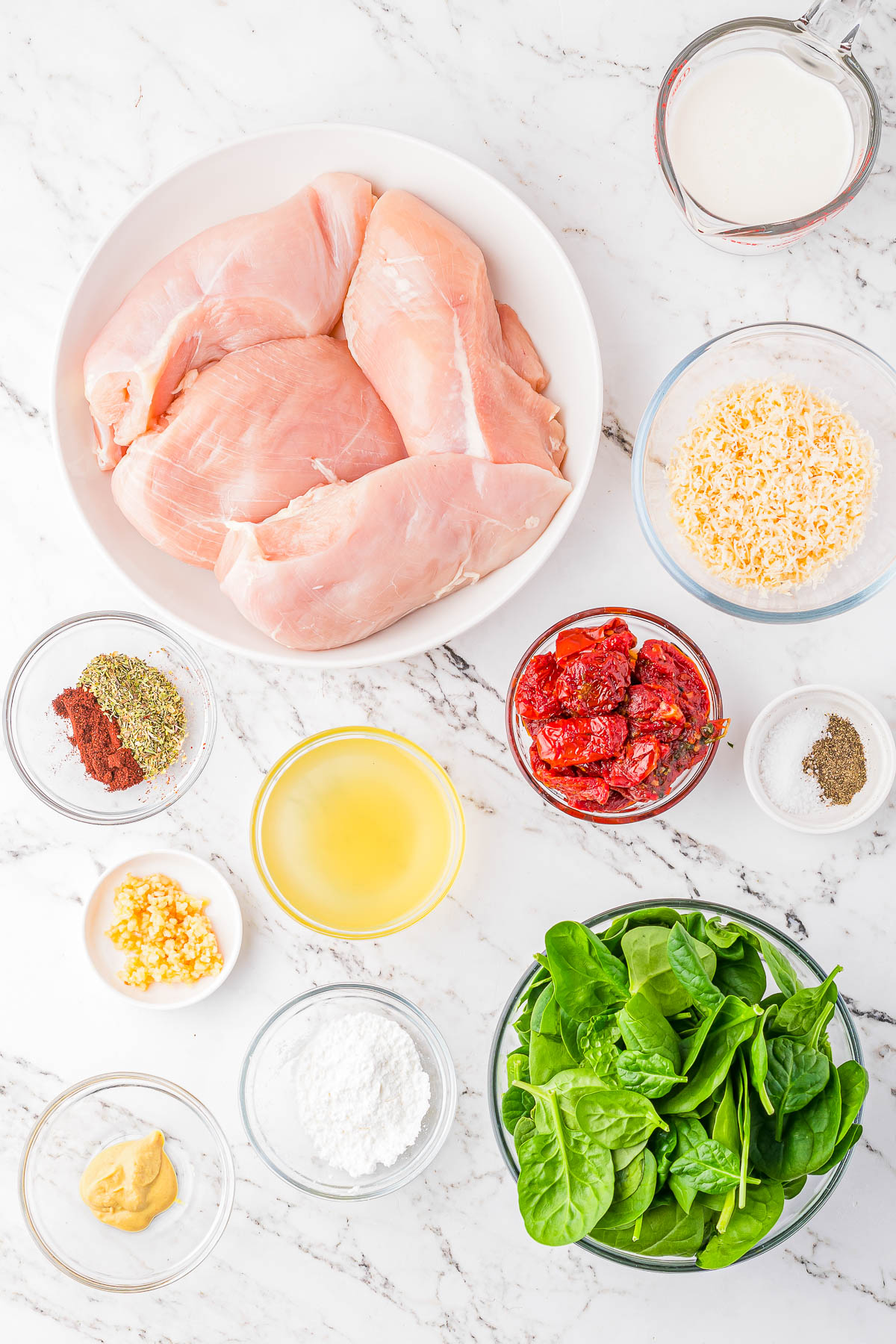 Various ingredients are arranged on a marble surface, including raw chicken breasts, fresh spinach, shredded cheese, sun-dried tomatoes, cream, garlic, spices, broth, mustard, and cornstarch.