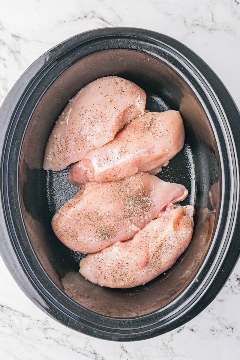 Four raw, seasoned chicken breasts placed inside a black slow cooker on a white marble countertop.