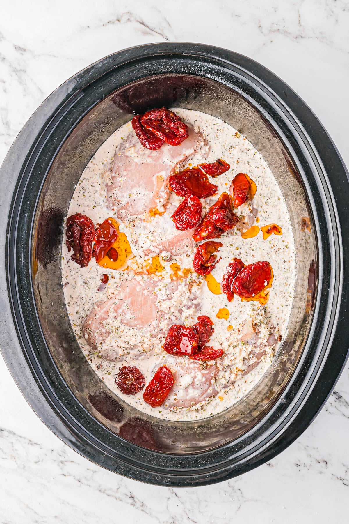 Raw chicken breasts, creamy white sauce with herbs, and sun-dried tomatoes in a slow cooker on a marble countertop.