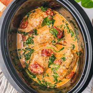 Chicken breasts cooked in a creamy sauce with spinach and sun-dried tomatoes in a slow cooker, with fresh spinach and parmesan cheese nearby.