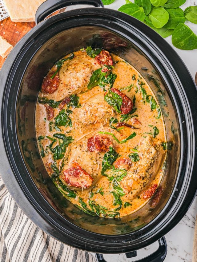 Chicken breasts cooked in a creamy sauce with spinach and sun-dried tomatoes in a slow cooker, with fresh spinach and parmesan cheese nearby.