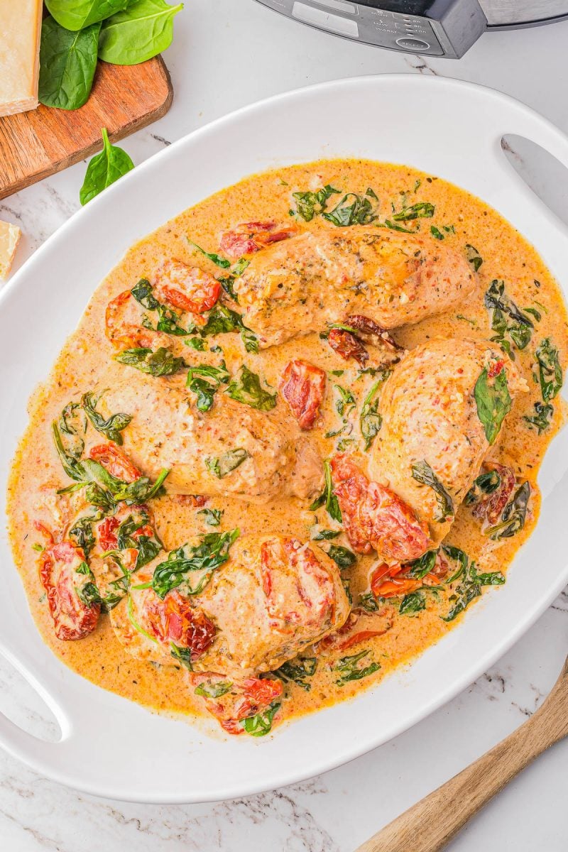 Four pieces of chicken in a creamy sauce with spinach and sun-dried tomatoes on a white oval dish.