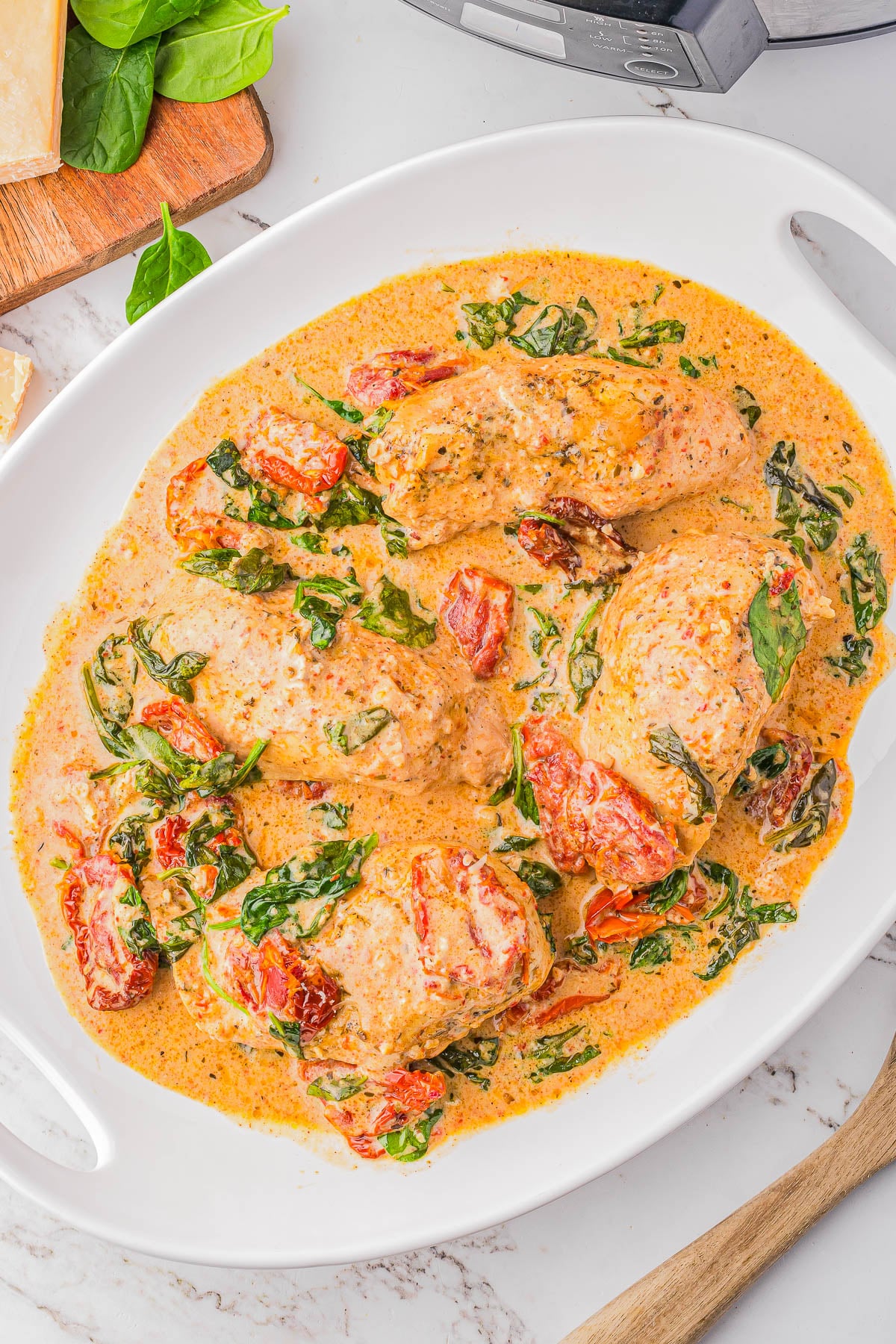 Four pieces of chicken in a creamy sauce with spinach and sun-dried tomatoes on a white oval dish.