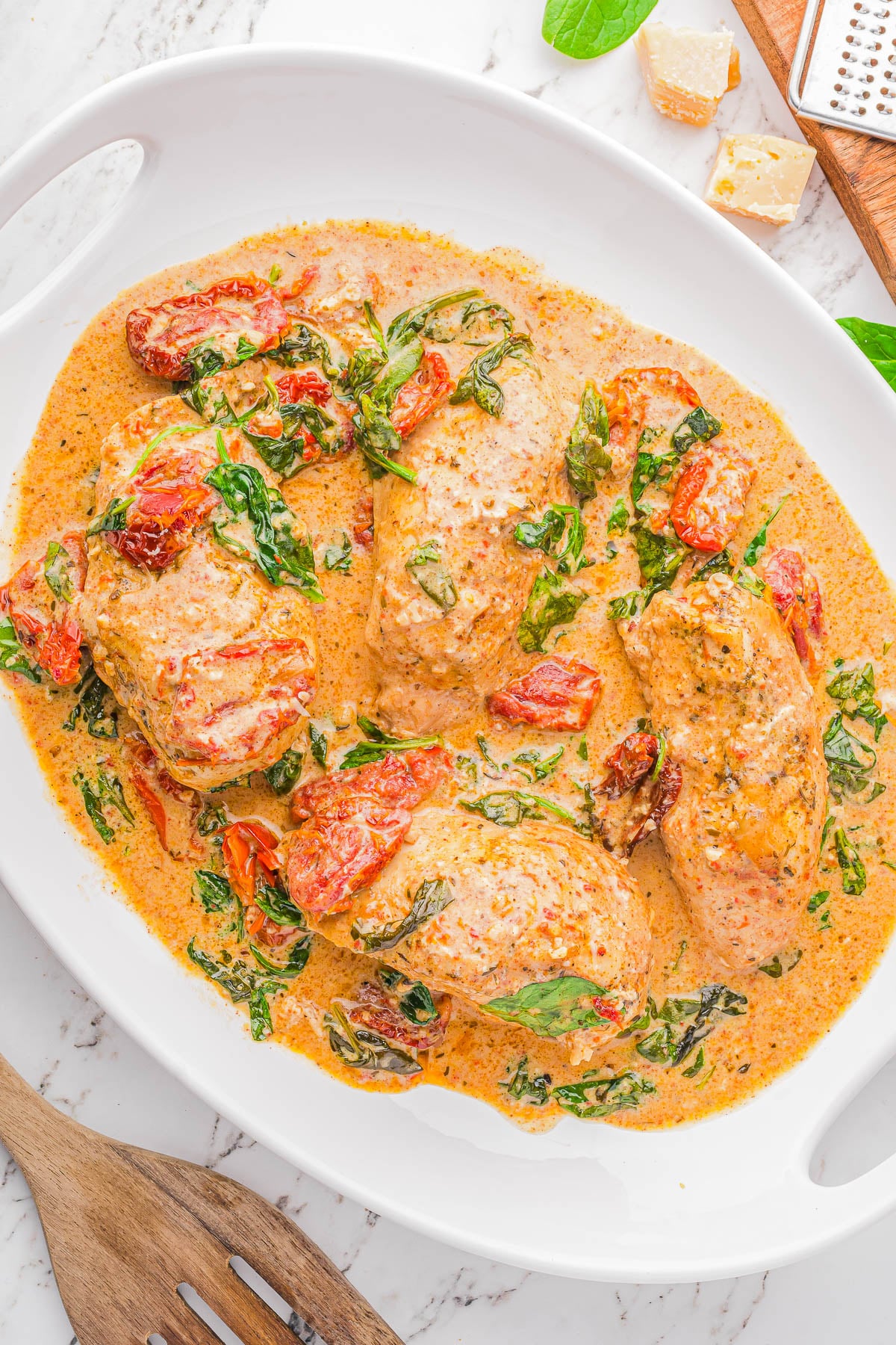 Four pieces of chicken breast cooked in a creamy sauce with spinach and sun-dried tomatoes, served in a white dish on a marble surface.