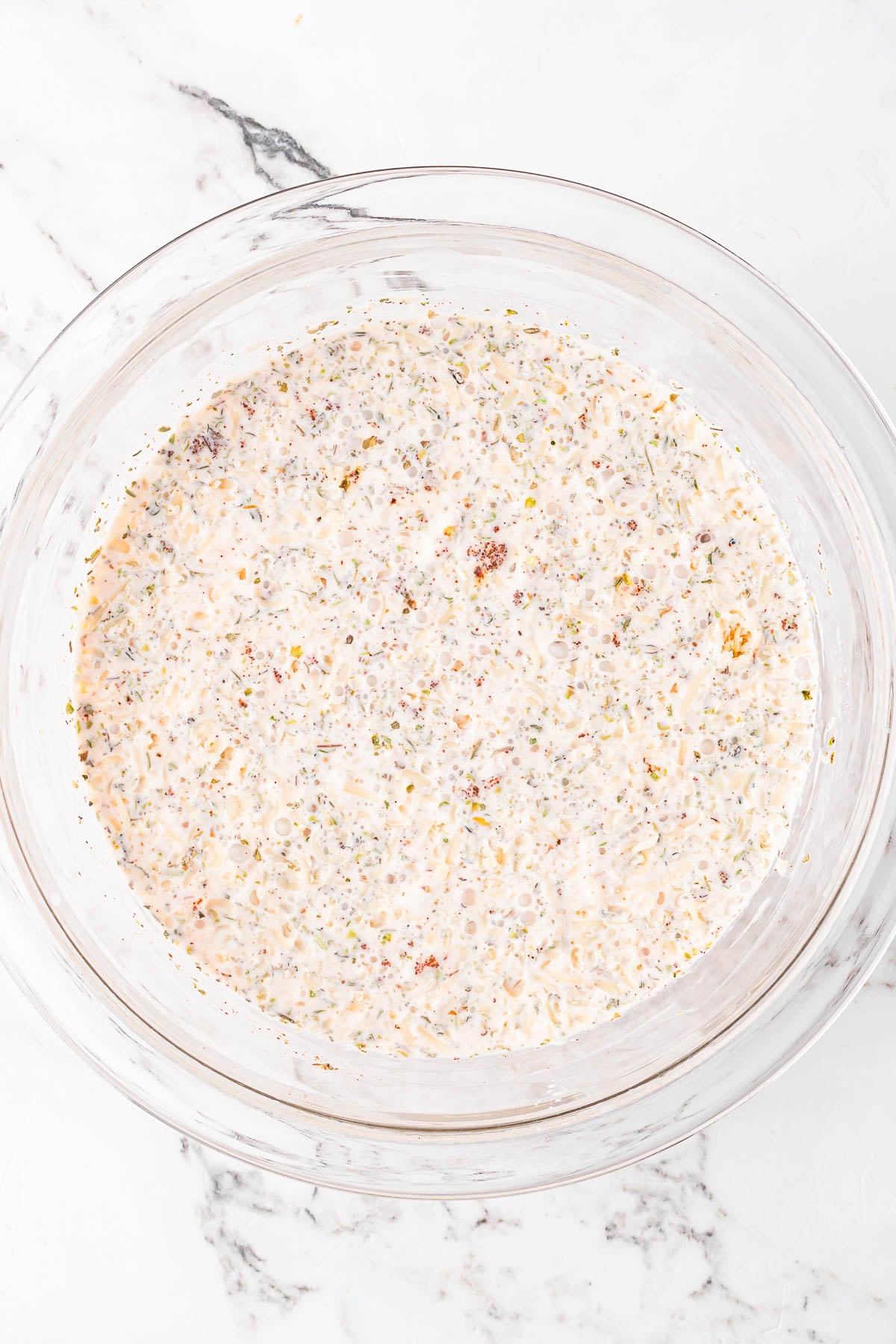 A clear glass bowl filled with a creamy, speckled mixture sitting on a white marble surface.