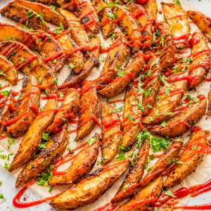 Seasoned potato wedges garnished with chopped herbs and drizzled with ketchup, arranged on parchment paper.