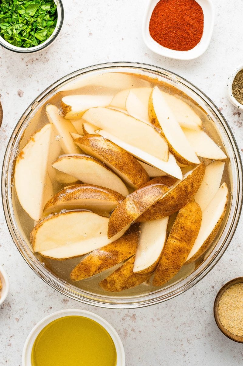 A glass bowl of potato wedges soaking in water, surrounded by small bowls containing chopped herbs, paprika, black pepper, olive oil, and other seasonings.