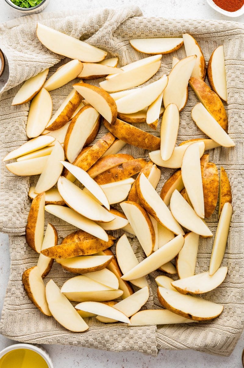 Potato wedges with skin on are spread out on a beige textured cloth, ready for cooking prep.