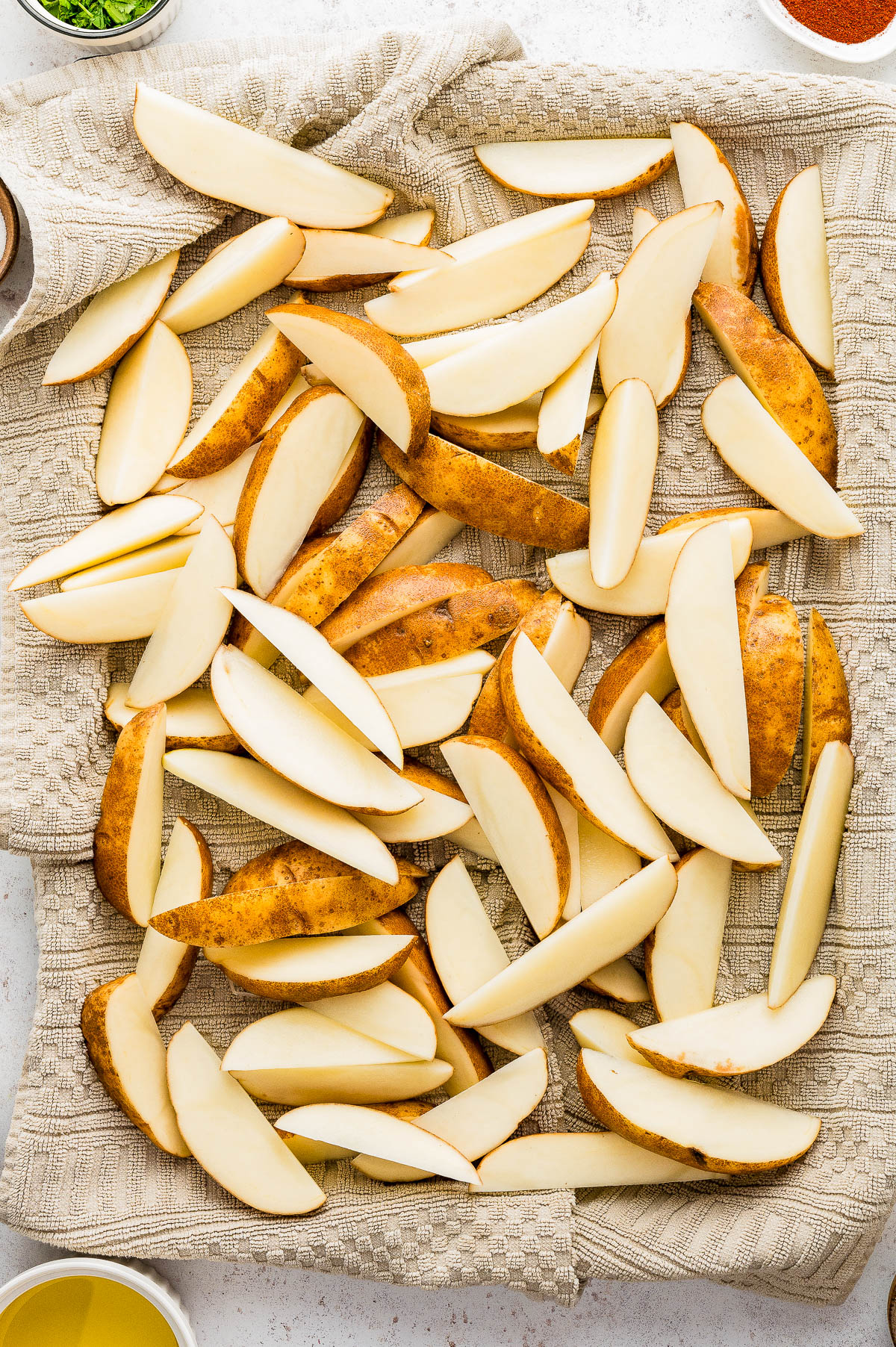 Potato wedges with skin on are spread out on a beige textured cloth, ready for cooking prep.