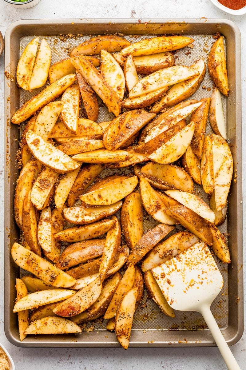 A baking sheet filled with seasoned potato wedges and a white spatula, ready for roasting.