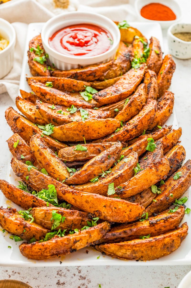 A plate of seasoned potato wedges garnished with chopped herbs, served with a small bowl of ketchup.