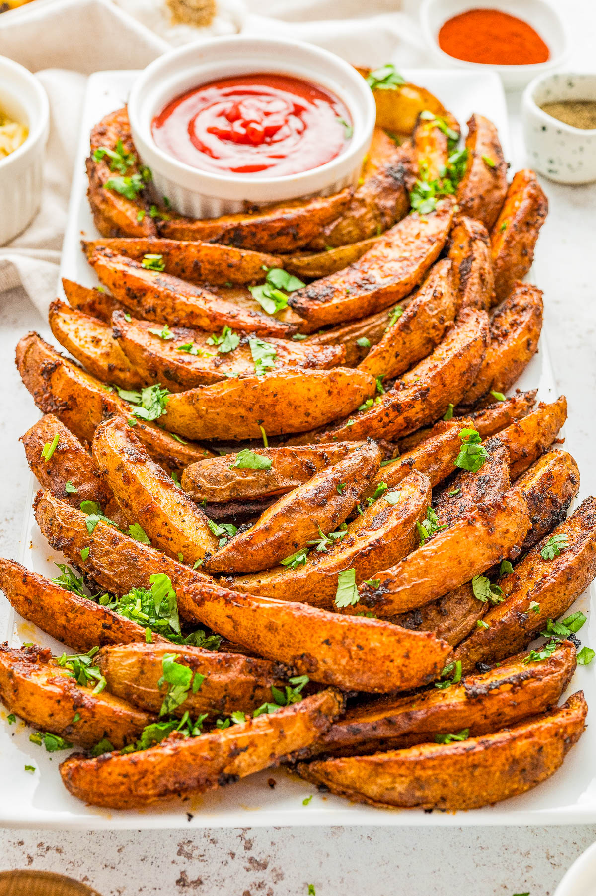 A plate of seasoned potato wedges garnished with chopped herbs, served with a small bowl of ketchup.